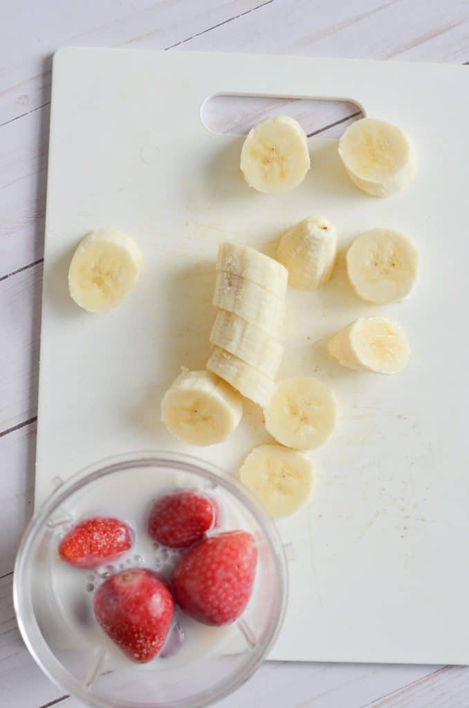 Smoothie ingredients in blender container and sliced bananas on counter.