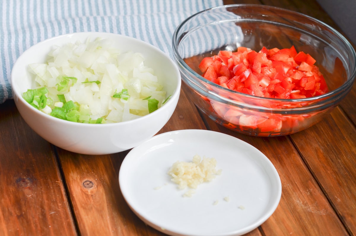 bowl of chopped red peppers, bowl of chopped onions and green onions and plate with chopped garlic.